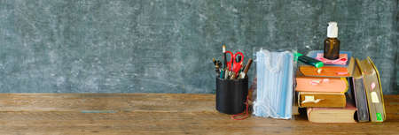 School supplies and COVID 19 prevention items on classroom desk with books, eyeglasses, pens on chalkboard background. Back to school during corona-virus pandemic concept.の写真素材