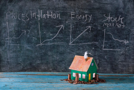 Rising rents, heating costs and prices for homes, model home with stacked money in front of blackboard with financial diagrams.Free copy spaceの写真素材