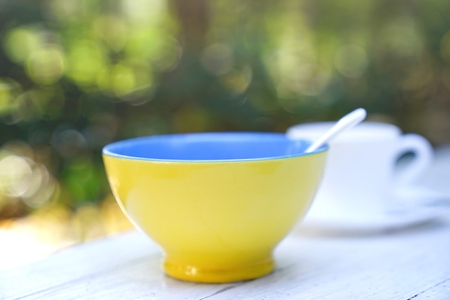Yellow blue bowl with white spoon and white coffee cup on wooden table. Breakfast in green garden.の写真素材