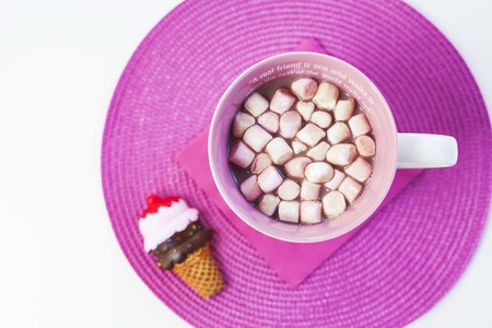 Cup of hot chocolate with sweet marshmallows on a pink placement with ice cream cone magnet. A...