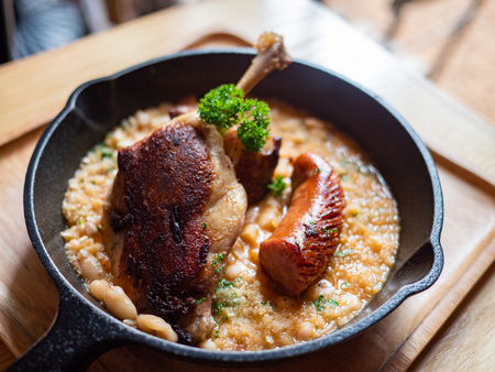 Sizzling hot, rich, slow-cooked Trio Cassoulet of duck confit, pork belly and smoked sausage on a bed of white beans in a black cast iron pan on a wooden table. Natural Light.の写真素材