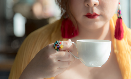Asian woman hold a cup of fresh brew tea or coffee during afternoon tea or high tea against herself wearing red lips with colorful fancy earring and ring. Elegant oriental setting. Natural lighting.の写真素材