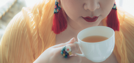 Asian woman hold a cup, smile to sip fresh brew tea during afternoon tea or high tea against herself wearing red lips with colorful fancy earring and ring. Elegant oriental setting. Natural lighting.の写真素材