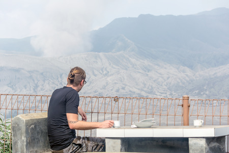 Mt. Bromo, A tourist looking at the Mount Bromo spewing volcanic  ashes from its, crater while drinking coffee.のeditorial素材