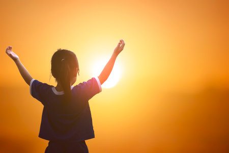 Silhouette of little girl raising hands on sunset sky background.の写真素材
