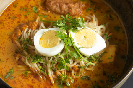 Laksa Noodle Soup rich with coconut milk topped with noodles, tofu puffs, fish cakes, bean sprouts, garnished with coriander and boiled eggs served in bowl on black background closeup side viewの写真素材