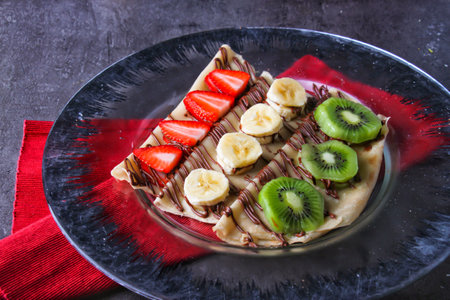 Colorful Fruit Crepe with Chocolate opped with fresh strawberries, bananas, and kiwi slices, with rich chocolate sauce served in plate isolated on table side view of indulgent dessertの写真素材