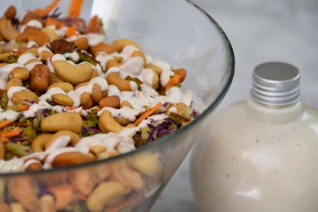 Fresh Broccoli Salad with Cashews in a clear glass bowl with broccoli florets, crunchy cashews, carrots, and red cabbage, drizzled with creamy white sauce bottle closeup view of healthy saladの写真素材
