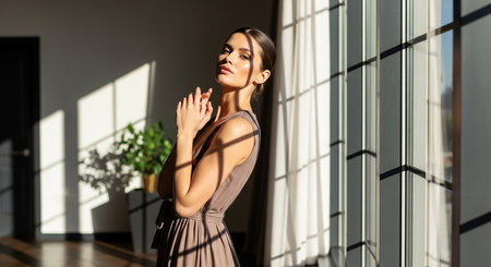 beautiful woman in evening dress looking away while standing near window at homeの素材