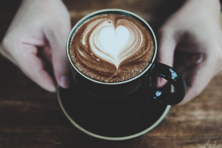 Hands holding cup of hot coffee with heart shaped foam, warm color tone , good service coffee shopの写真素材
