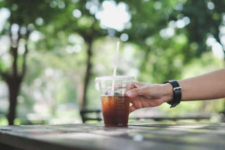 man hand touching a plastic cup of iced americano (black coffee) on the wood plank table in the gardenの写真素材