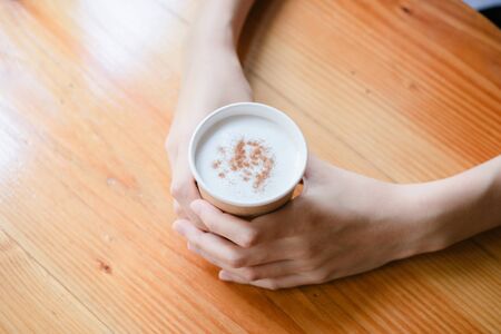 Hot drink in a craft plastic cup with a white cap in the hands of a young girl with a red manicure in a black coat close-up on the street in the fall afternoon.の写真素材
