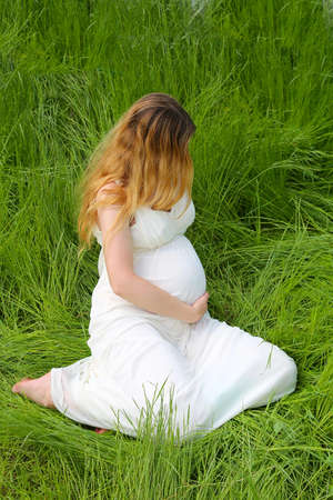 A pregnant girl at a late date in a white dress is sitting on the grass, hugging her stomachの写真素材