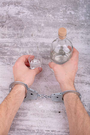 The concept of male alcohol addiction. Men's handcuffed hands hold a bottle and a glass of alcohol.の写真素材