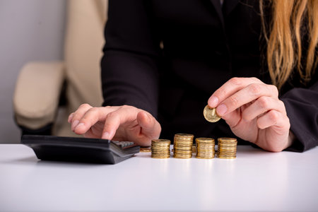 A business woman counts on a calculator and holds a coin in her hand, at a table, in front of stacks of coins, close-upの写真素材