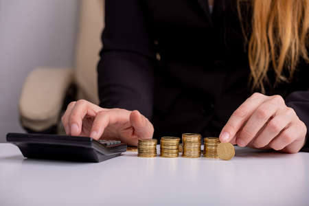A business woman counts on a calculator and holds a coin in her hand, at a table, in front of stacks of coins, close-upの写真素材