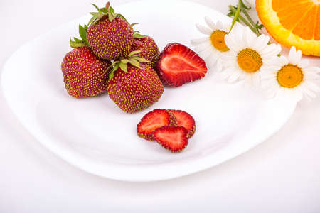 Ripe strawberries, whole and in pieces, on a white plate against a background of daisies and juicy orange, on a light background, close-upの写真素材