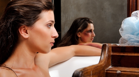 Woman relaxing in bathtub with mirror image of her with bruises on her face, a conceptual shoot of...