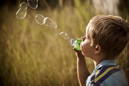Young blonde boy blows a long string of bubbles in a field; image graded in a vintage tone 