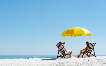 Beach summer couple on island vacation holiday relax in the sun on their deck chairs under a yellow umbrella. Idyllic travel background.の販売画像