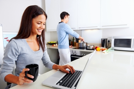 woman working on laptop in kitchen as boyfriend prepares meal. happy healthy relationship multiracial coupleの写真素材