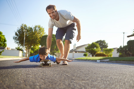 young boy learning to ride skateboard as father teaches him in the suburb street having fun.の写真素材