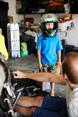 portrait of young boy playing with fathers motorbike helmet and helping his dad with fixing a motorcycle in the garageの写真素材