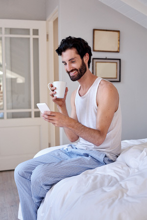 man with mobile cellphone and coffee sitting on bed at homeの写真素材