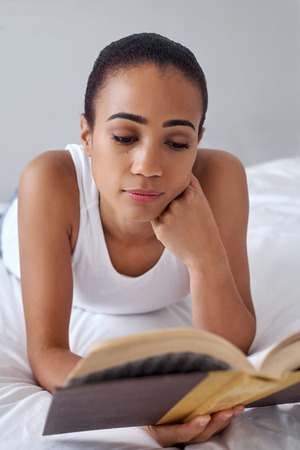 young woman laying relaxing on bed reading literature novel story book at home
