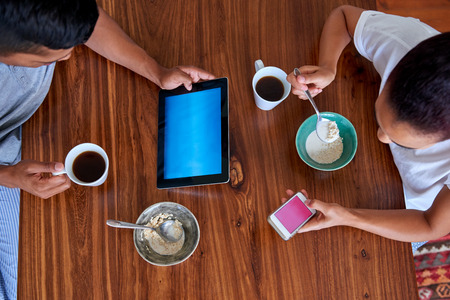 couple enjoying a healthy morning breakfast cereal with tablet computer and mobile phone in kitchen at homeの写真素材
