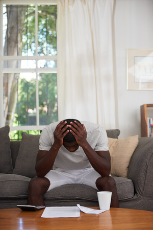 disapointed troubled african black man sitting on sofa couch calculating home bill finance debt in living roomの写真素材