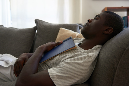 african black man sleeping on sofa couch while reading book at home living room loungeの写真素材