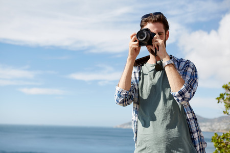 young, attractive man standing and taking pics of the ocean using a digital cameraの写真素材
