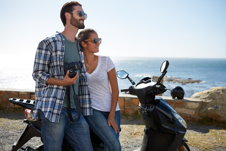 happy couple standing against a scooter, enjoying the beautiful ocean viewの写真素材