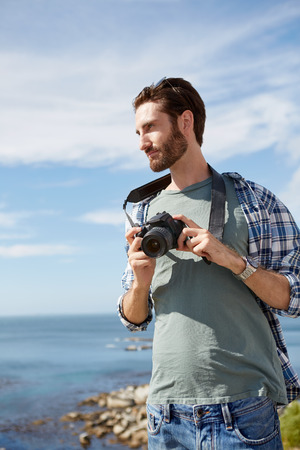 young, attractive man stands near the ocean with digital camera in his handの写真素材