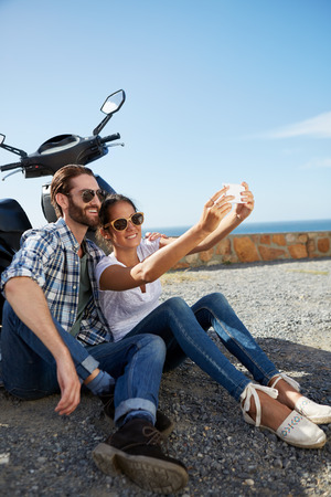 happy couple sitting near the ocean, against their scooter, taking a selfie on holidayの写真素材
