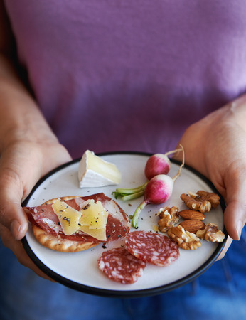 Hands holding a glass of wine and a plate of snacks of cured meat nuts fruit and finger foodの写真素材