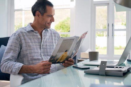 Casual businessman seated at home office space reading business magazine smiling