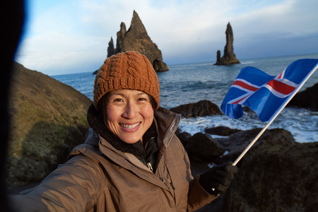 happu independent woman traveller at black sand beach south iceland, taking a selfie at reynisdrangar stone sea stacks, waving icelandic flagの写真素材