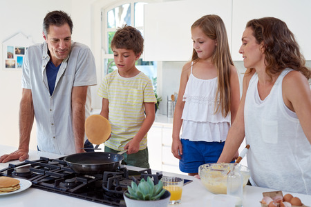 Happy caucasian family standing around stove, son making pancakes on stoveの写真素材