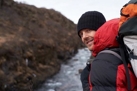 Smiling portrait of a man hiking in winter with beanie jacket and backpack on cliff overlooking a riverの写真素材
