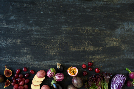 Border of fresh purple toned vegetables and fruits on dark rustic distressed background, eggplant, carrot, fig, aubergine, grapes, radishes, leaf lettuce, passionfruit, cherriesの写真素材