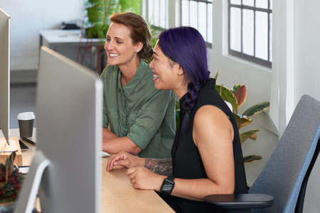Female coworkers having a discussion at the computer and laughing together, engaging and friendly colleaguesの写真素材