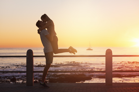 Young woman jumps on boyfriend and gives him a kiss at sunset, romantic passionate moment on date, golden sun flareの写真素材
