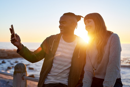 Multi racial friends couple smiling for cellphone selfie outdoors at sunset by oceanの写真素材