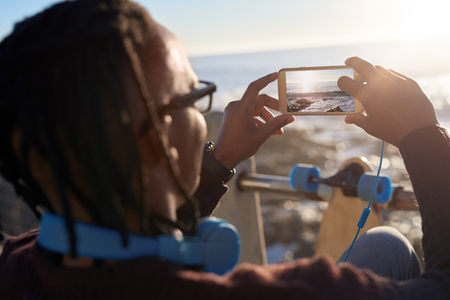 Trendy young man with dreadlocks taking photo of sunset on his mobile cell phone, beautiful scenary while skateboardingの写真素材