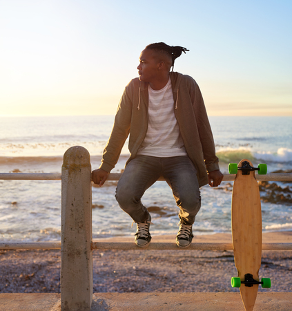 Young mixed race man sitting on railing with longboard, enjoying sunset on horizonの写真素材