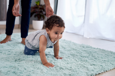 Baby boy crawling on rug carpet with mother parent in background helping, involved parentingの写真素材
