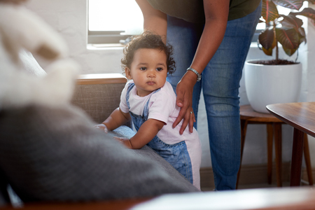 Mixed race baby girl cruising along sofa couch supported by mom, learning how to walk move growing developement skillの写真素材