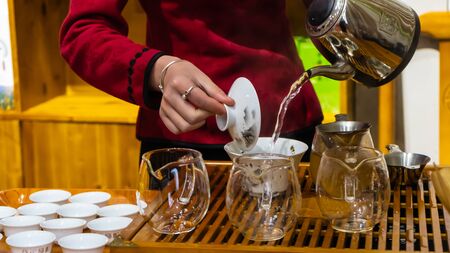 Close up shot at the hands of young Chinese woman wearing red dress and general chinese-styled bracelet and ring who is standing at the wooden tea station and making Chinese tea by holding a stainless steel teapot with one hand and the other hand holding tea lid and pouring hot water into a big chinese -styled white tea cup in order to split the tea into more than 10 small Chinese-styled white tea cups in the tea shopの写真素材